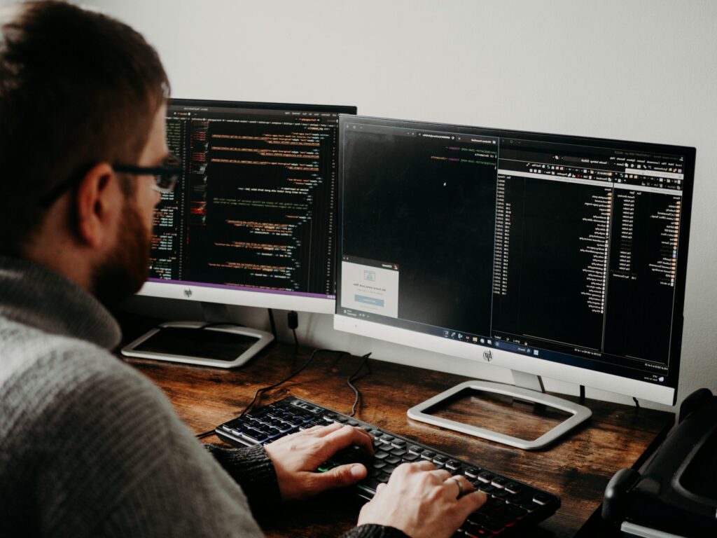 pexels photo 16129724 16129724 Software developer typing code on dual monitors at a wooden desk.